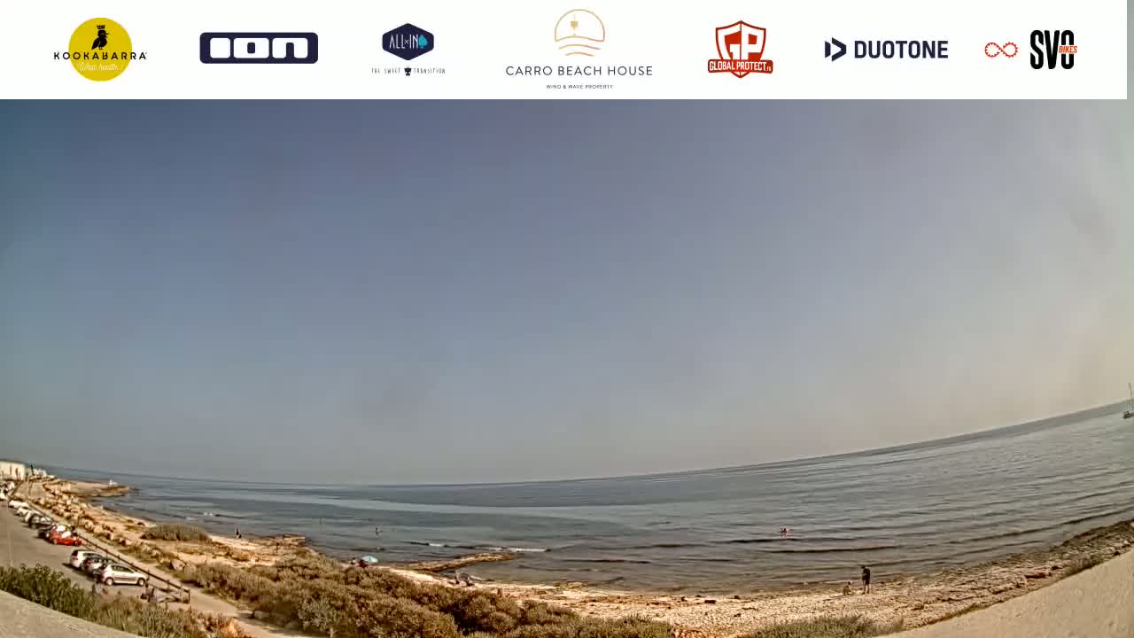 A wide, panoramic view of a calm ocean shore on a mostly clear day, with a sandy beach and a few people visible.