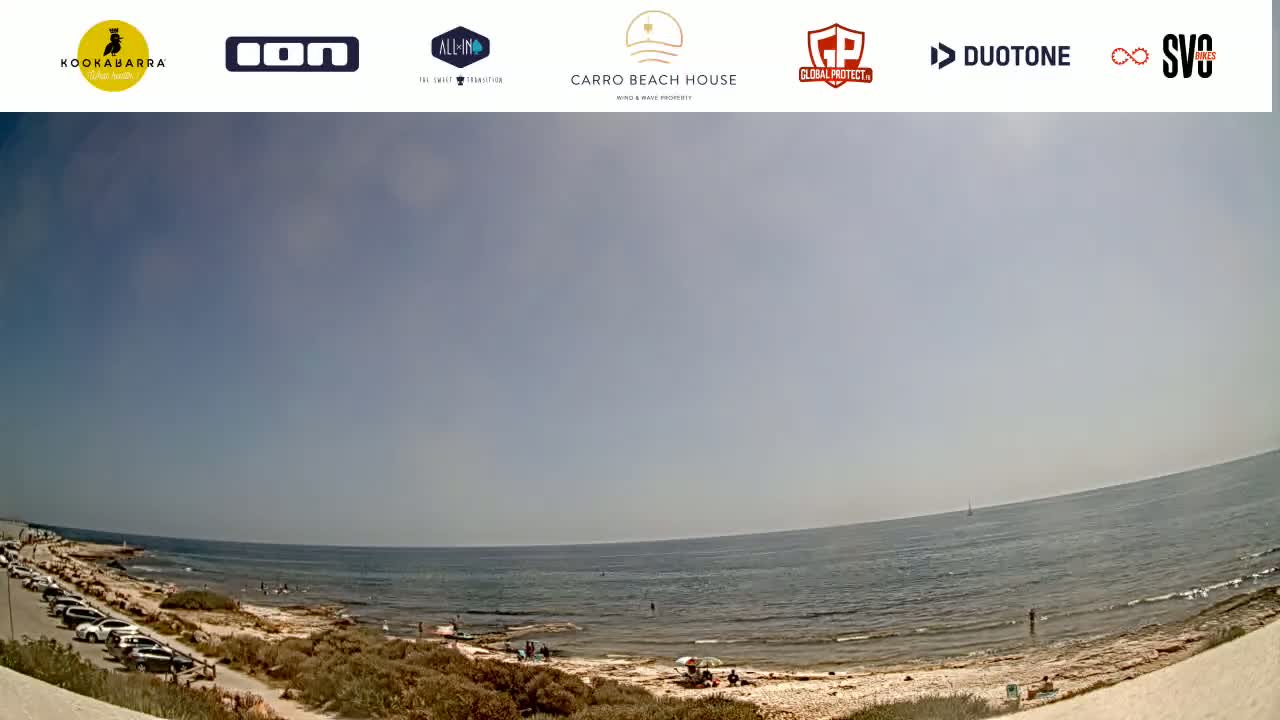 A mostly sunny day at a beach with calm ocean water, a sandy shore, and cars parked along a road above the beach.