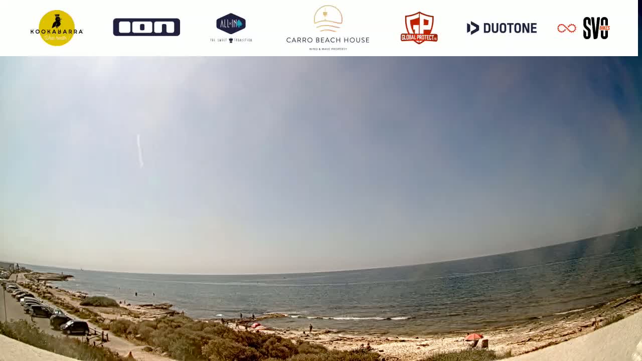 A mostly sunny day at a beach with calm water, a sandy shoreline, sparse vegetation, and a few people visible.