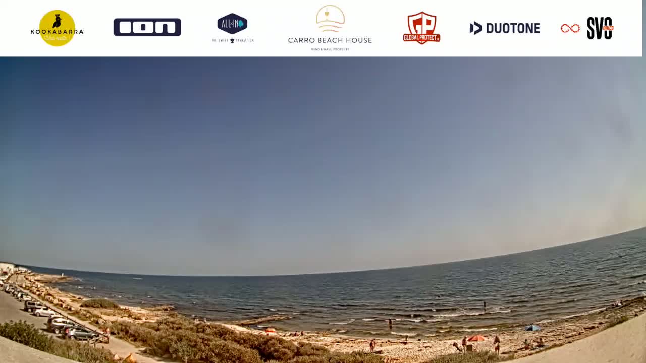 A mostly sunny day at a beach with calm water, a sandy shore, and a few people present.