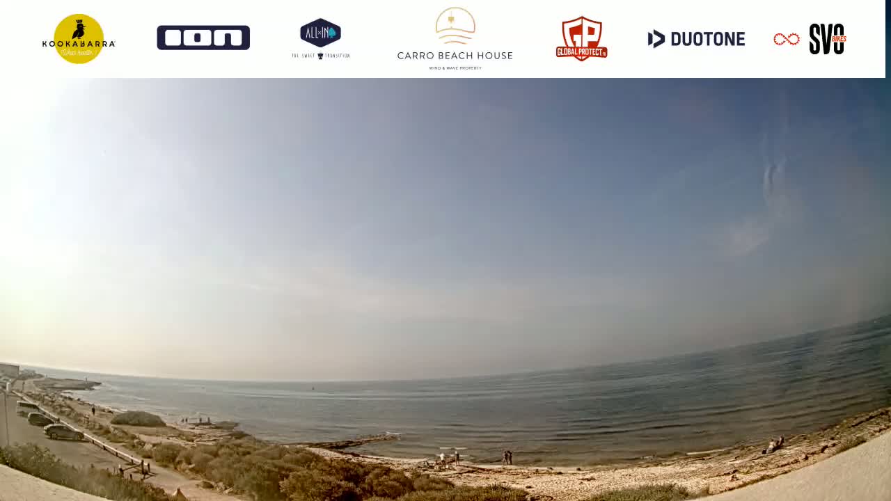 A mostly sunny day at a beach with calm ocean water, showing a sandy shoreline with sparse vegetation and a few people visible.