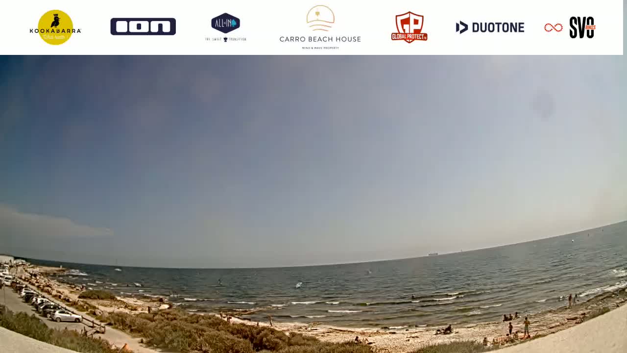 A mostly sunny day at a beach with calm waves, showing a sandy shore, some people, and a parking lot.