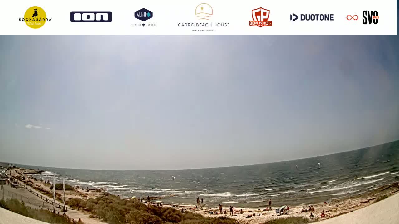 A mostly sunny day at a beach shows a wide expanse of ocean with small waves, a sandy shore with scattered people, and some vegetation along the coastline.
