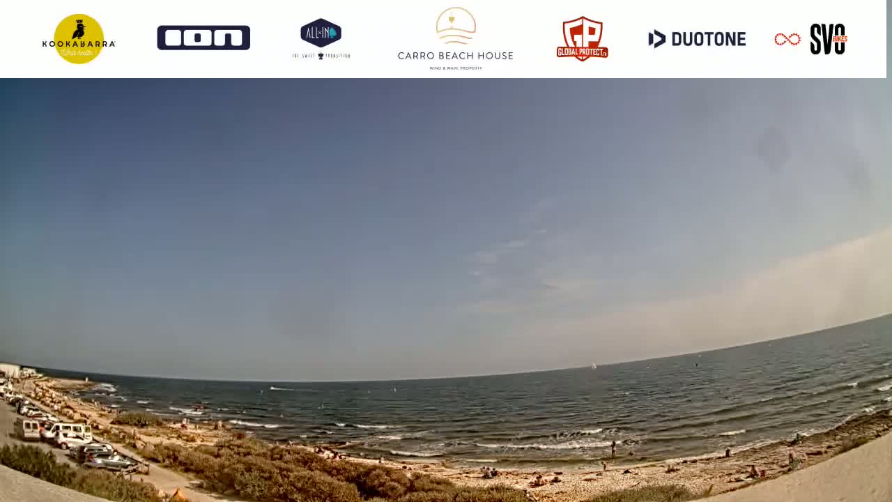 A mostly sunny day at a beach shows a calm sea with people swimming and relaxing on the shore, with a parking lot and vegetation visible in the foreground.