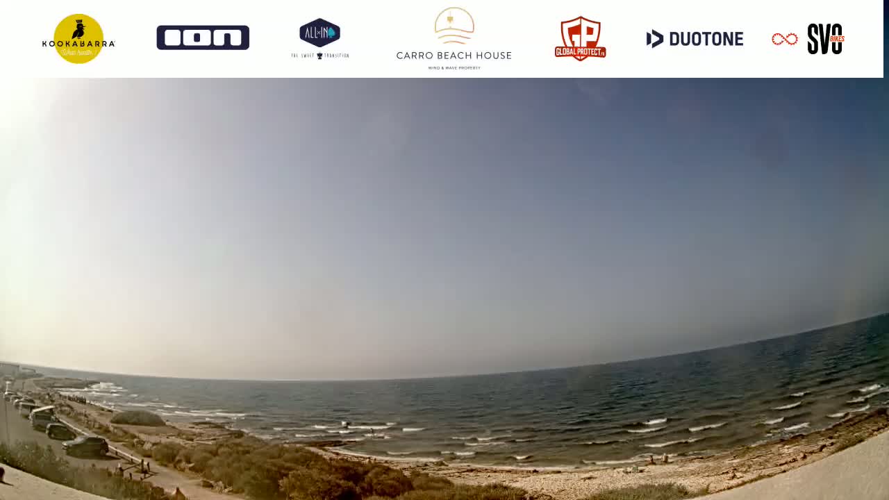 A wide, sunny shot of a calm ocean meeting a sandy shoreline with some people visible along the beach and a road.