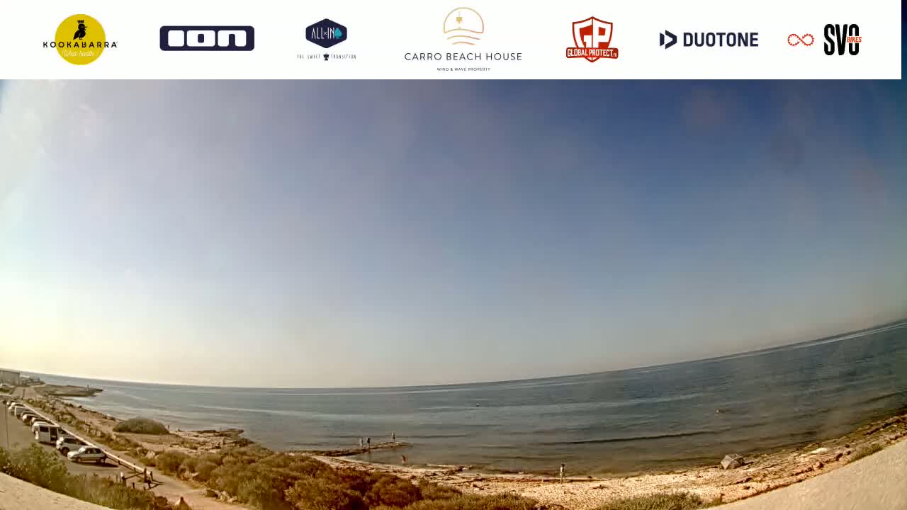 A wide, panoramic view shows a calm ocean under a clear, bright blue sky; a sandy shoreline and sparse vegetation are visible in the foreground.