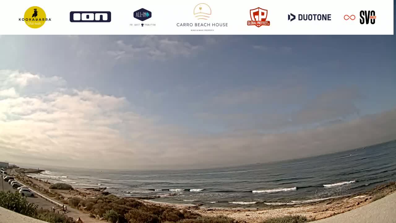 A mostly sunny day at a beach with small waves, a sandy shore, and a few people visible in the distance.