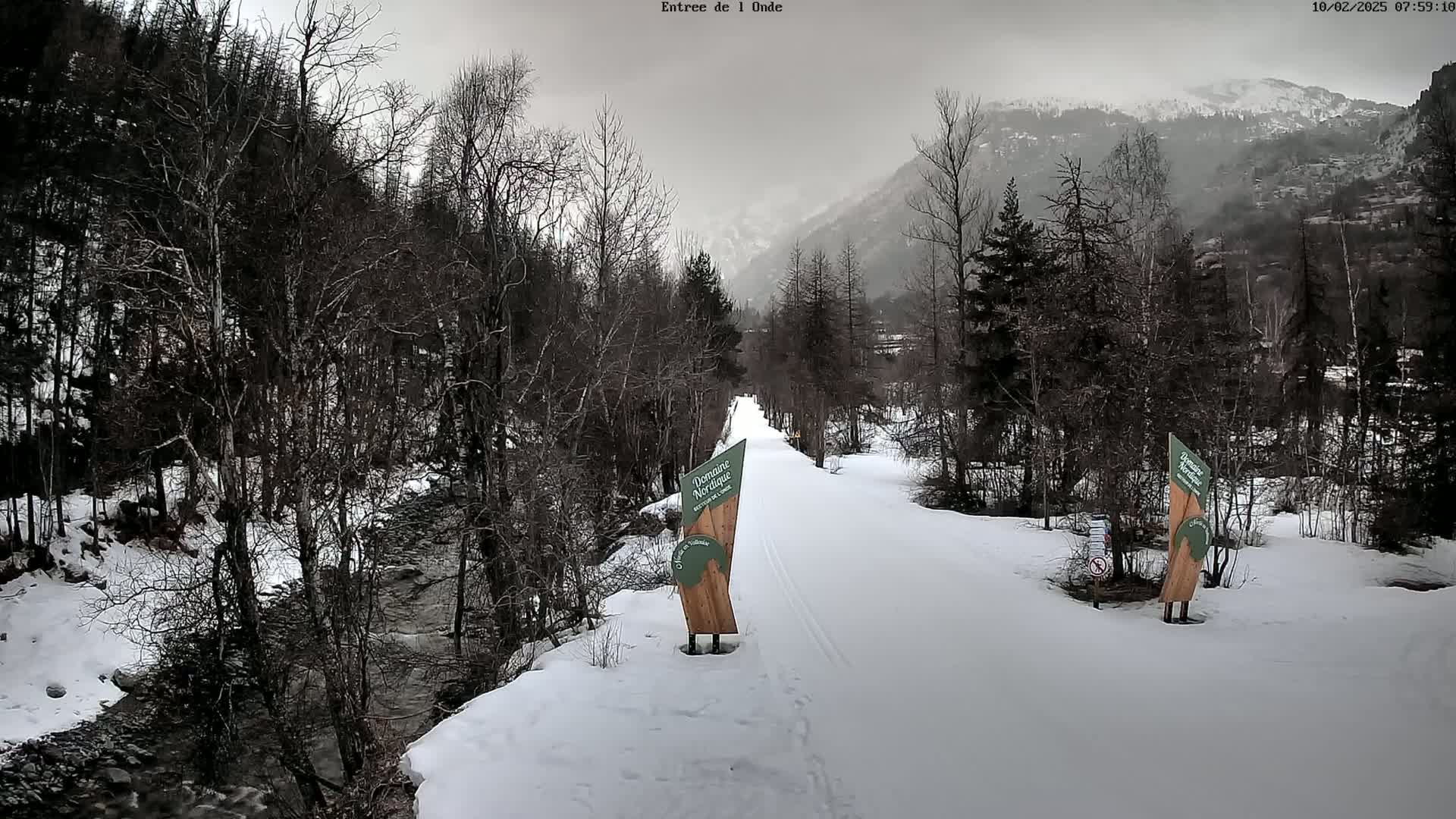 A snow-covered path winds through a snowy forest alongside a stream, with overcast skies and mountains in the background.
