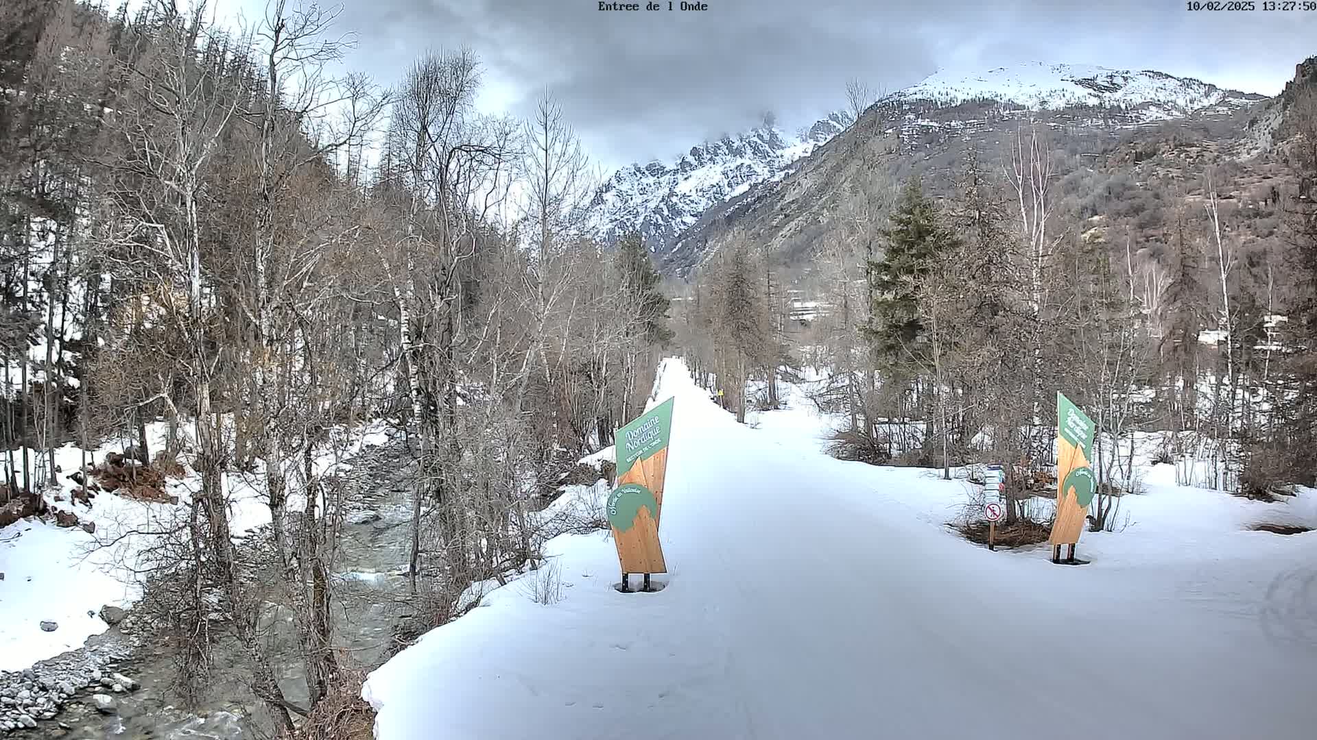 A snow-covered path winds through a valley of leafless trees alongside a flowing river, with snow-capped mountains in the background under an overcast sky.