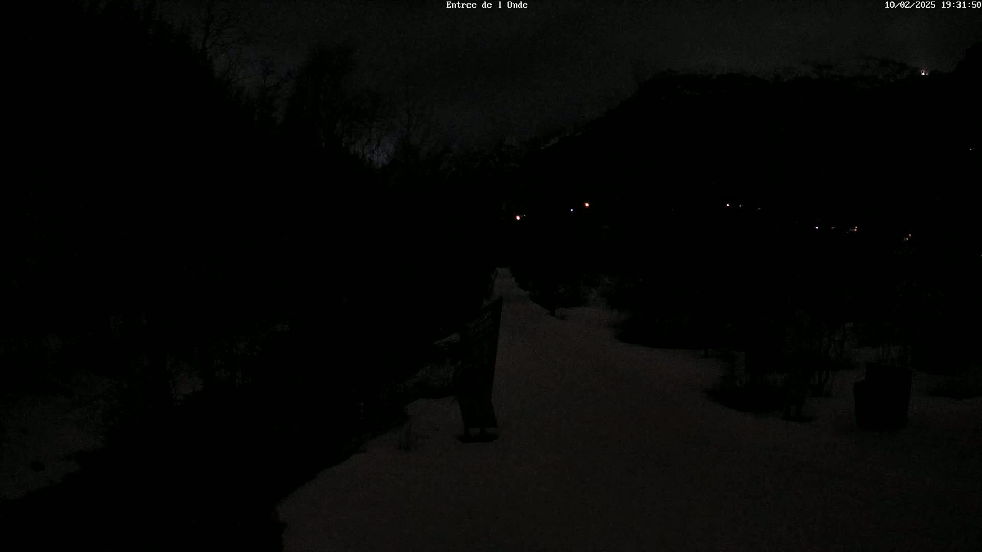 A dark, snowy path winds through a nighttime landscape of trees and distant lights under a cloudy sky.