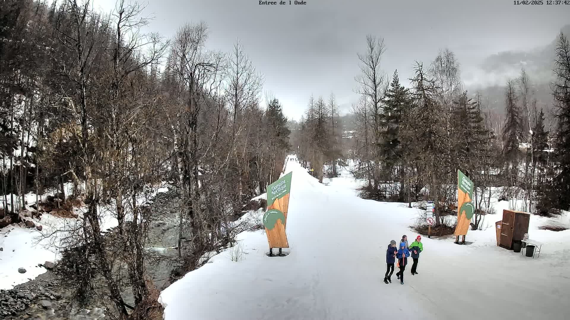 A snowy, overcast scene shows a snow-covered path winding through a wooded area alongside a partially frozen river, with a small group of people walking along the path.