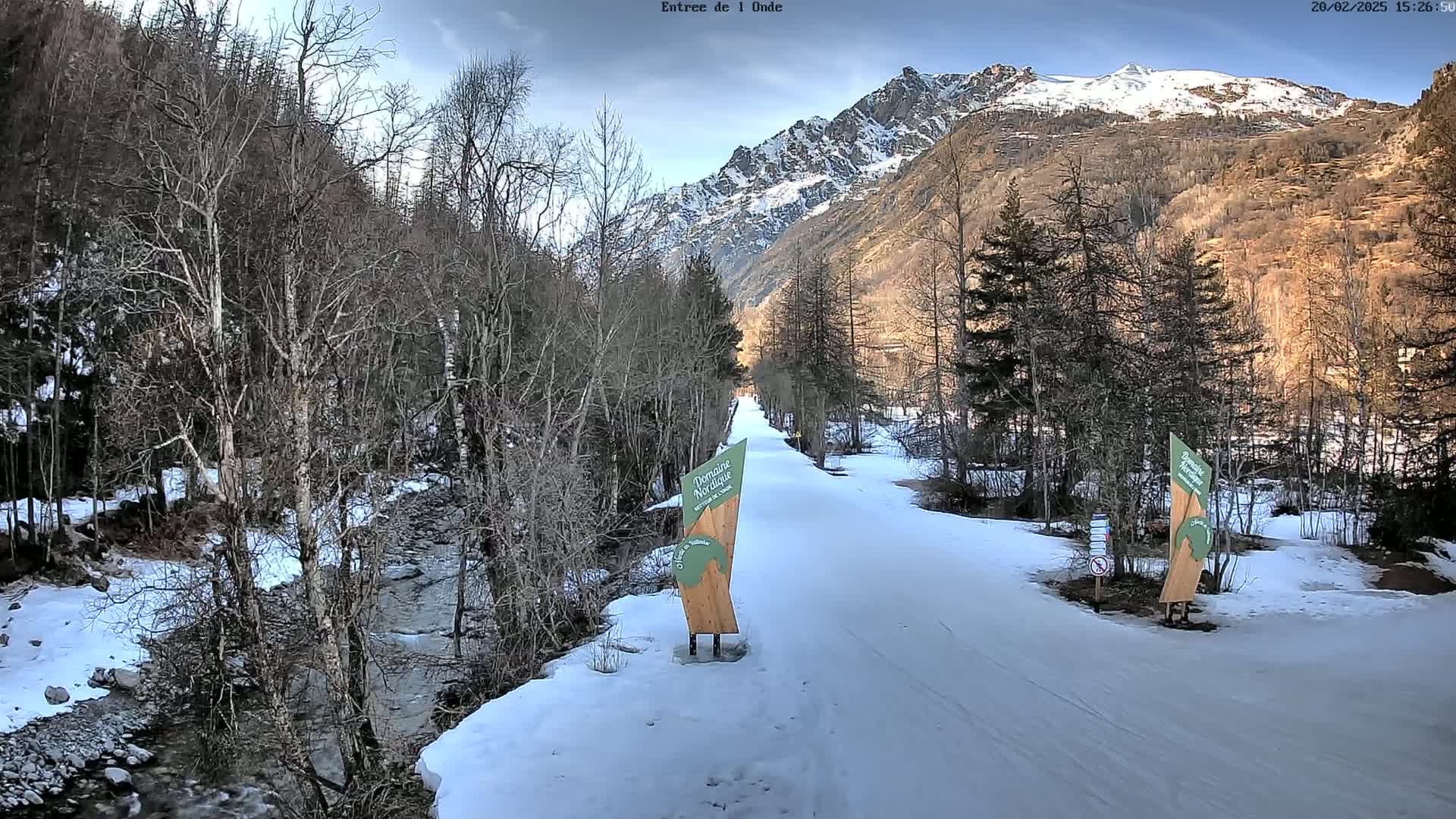 A snow-covered path winds through a valley of leafless trees and a small stream, with snow-capped mountains in the background under a partly cloudy sky.