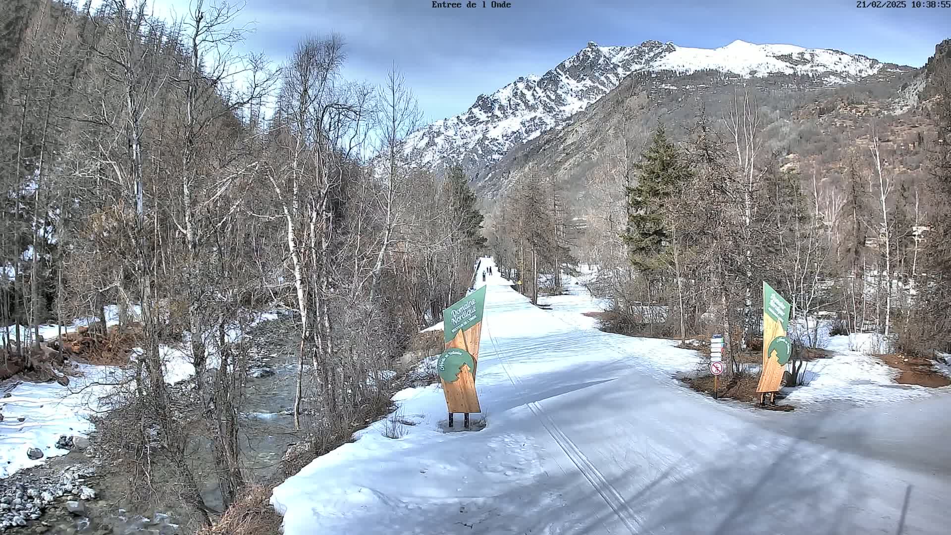 A snow-covered path winds through a valley of leafless trees and a partially snow-covered stream, leading toward a snow-capped mountain range under a partly cloudy sky.