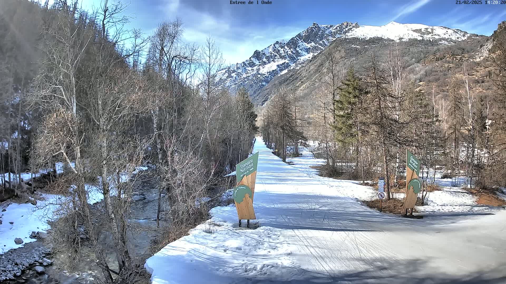 A snow-covered path winds through a valley of leafless trees alongside a stream, with snow-capped mountains in the background under a mostly sunny sky.