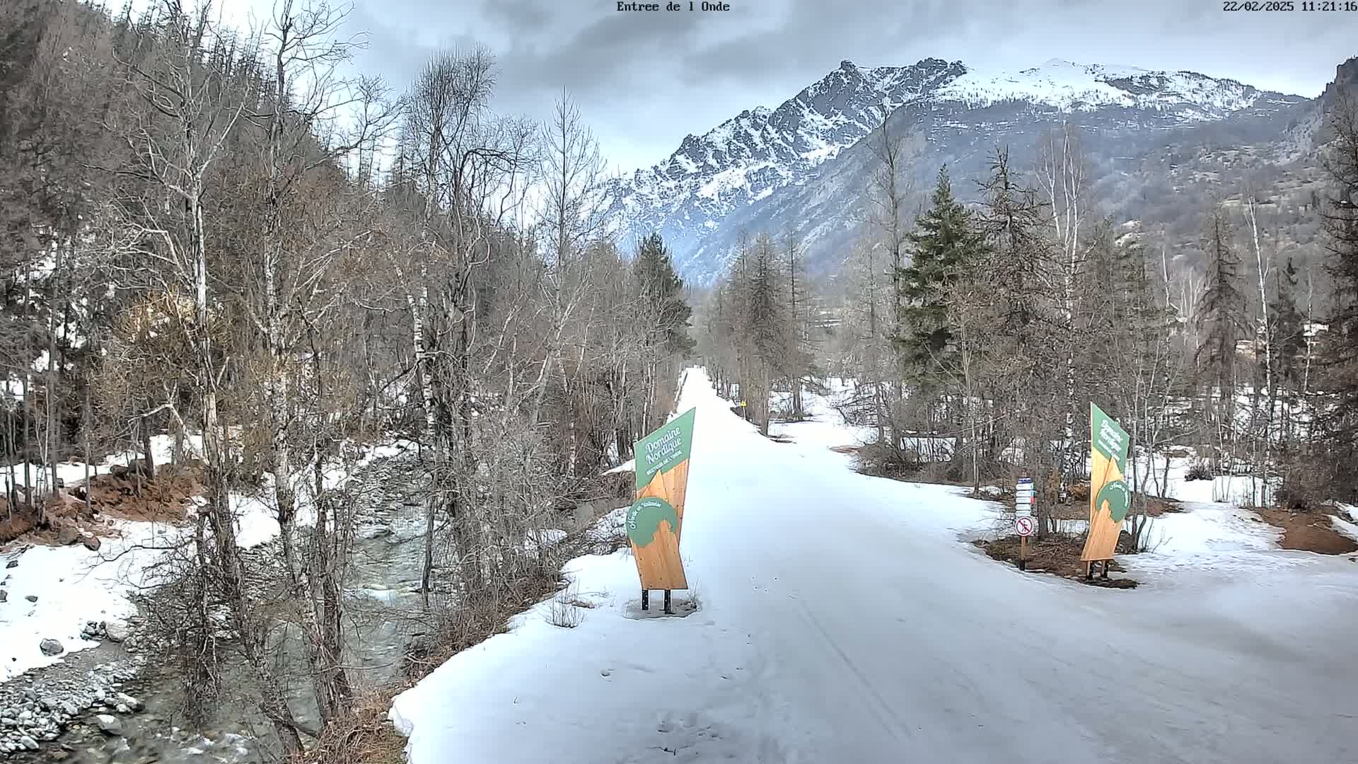 A snow-covered path winds through a valley, bordered by leafless trees and a flowing stream, with snow-capped mountains in the background under an overcast sky.