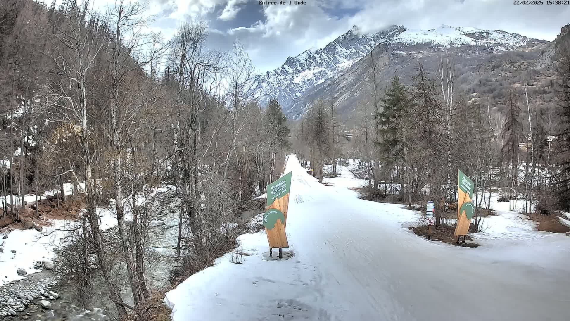 A snow-covered path winds through a valley of leafless trees beside a partially frozen stream, with a mountain range in the background under partly cloudy skies.