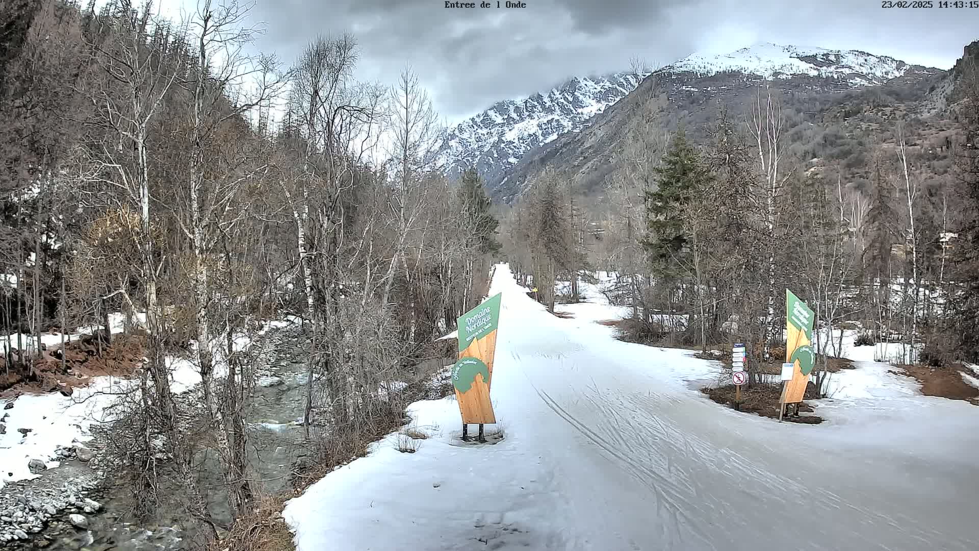 A snow-covered cross-country ski trail winds through a valley bordered by leafless trees and a snow-dusted mountain under an overcast sky.