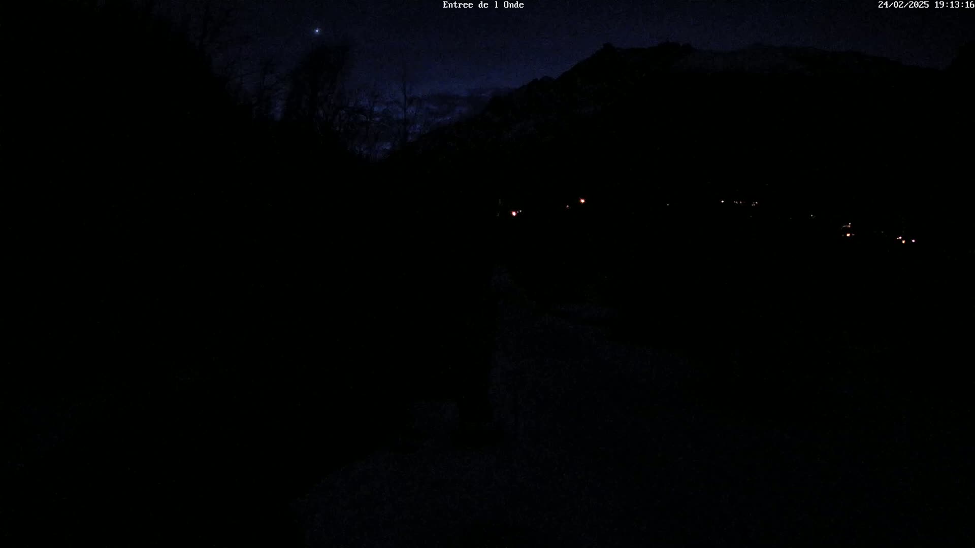 A nighttime, dark view of a road leading towards a mountain range with a few faint lights visible in the distance under a mostly clear, dark sky.