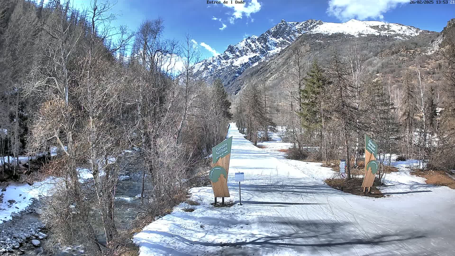 A snow-covered path winds through a valley, flanked by leafless trees and a partially snow-capped mountain range under a mostly sunny sky.