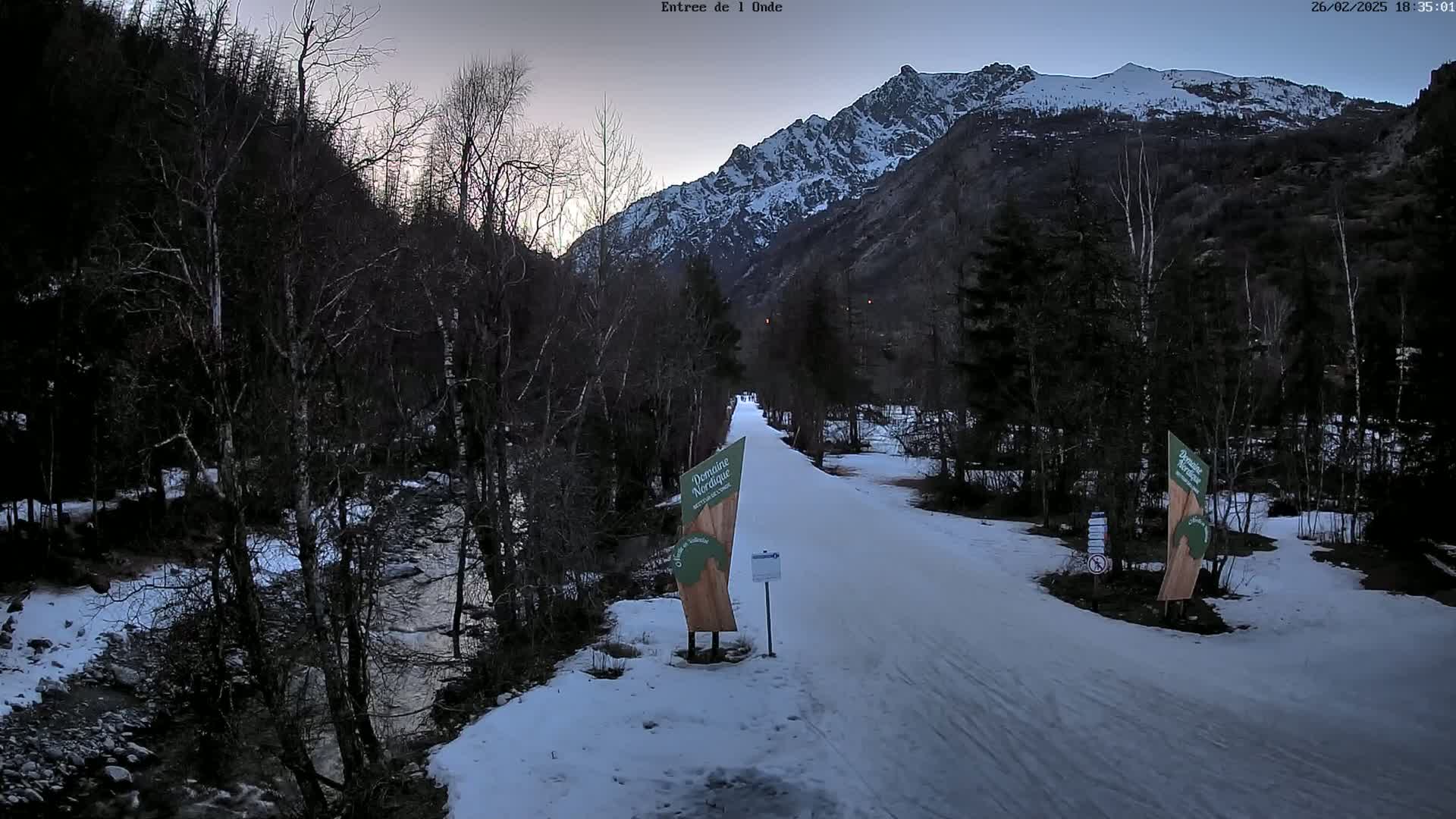 A snow-covered path winds through a valley beside a stream, with leafless trees and a snow-capped mountain range in the background under a twilight sky.