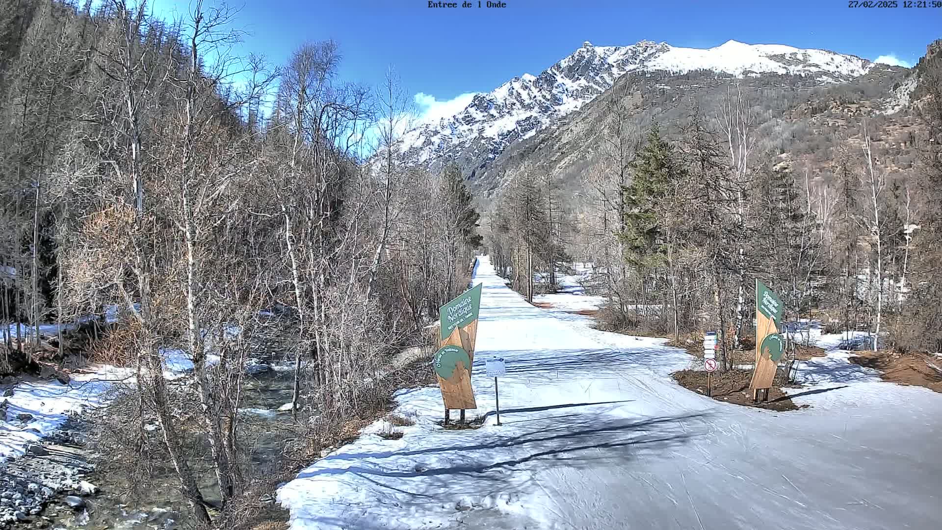 A snow-covered path winds through a valley bordered by leafless trees and a snow-capped mountain range under a clear blue sky.