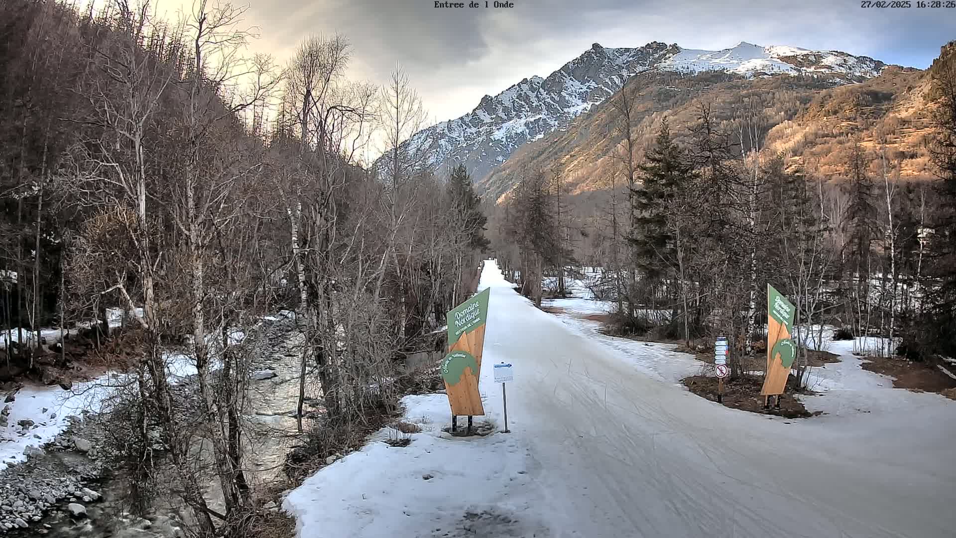 A snow-covered road winds through a valley bordered by leafless trees and a mountain range under a cloudy sky.