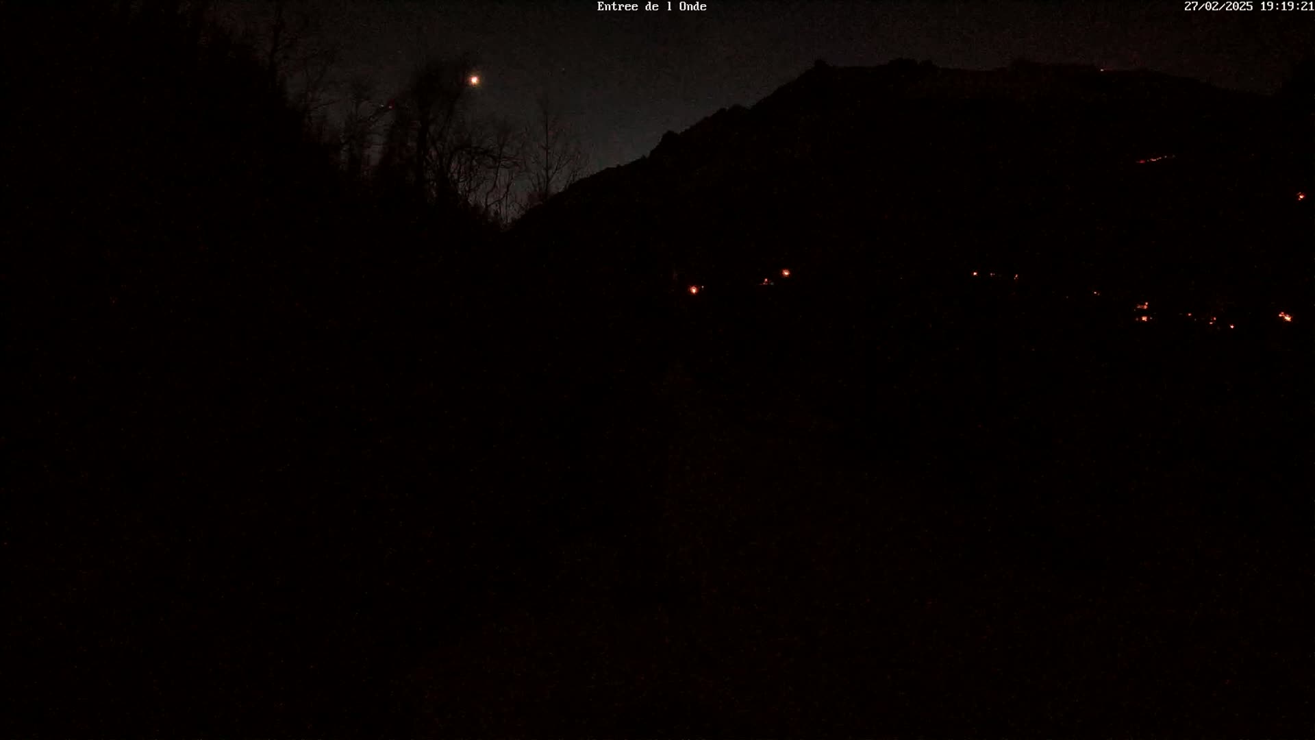 A nighttime, dark scene shows a silhouetted mountain and trees under a mostly clear sky with a bright moon visible.