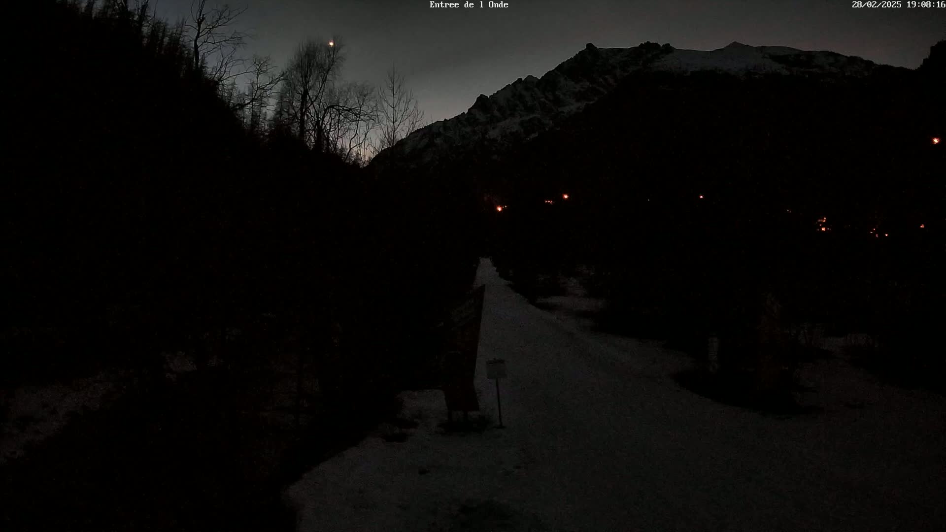 A dark, snow-covered path winds through a valley under a twilight sky, with a mountain range visible in the background and a few faint lights in the distance.