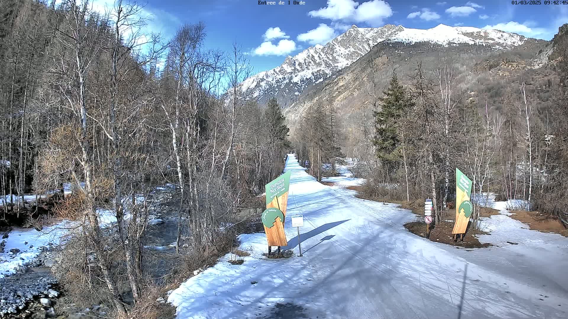 A snow-covered path winds through a valley of leafless trees toward a snow-capped mountain range under a partly cloudy sky.