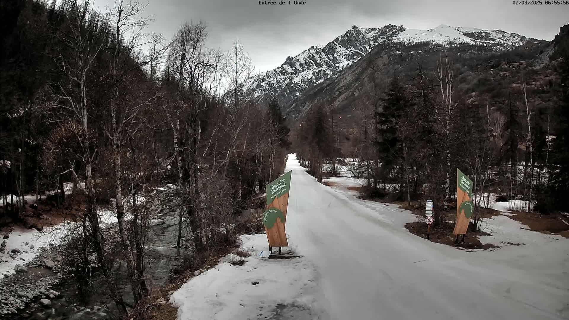 A snow-covered road winds through a valley of leafless trees and a snow-capped mountain under an overcast sky, with a small stream visible to the left.