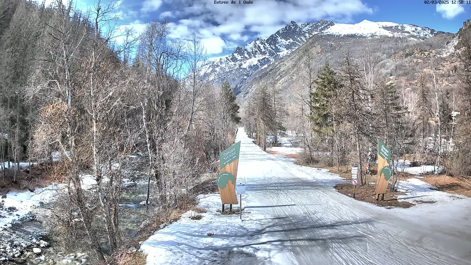 A snow-covered path winds through a valley of leafless trees and a partially snow-capped mountain range under a partly sunny sky.