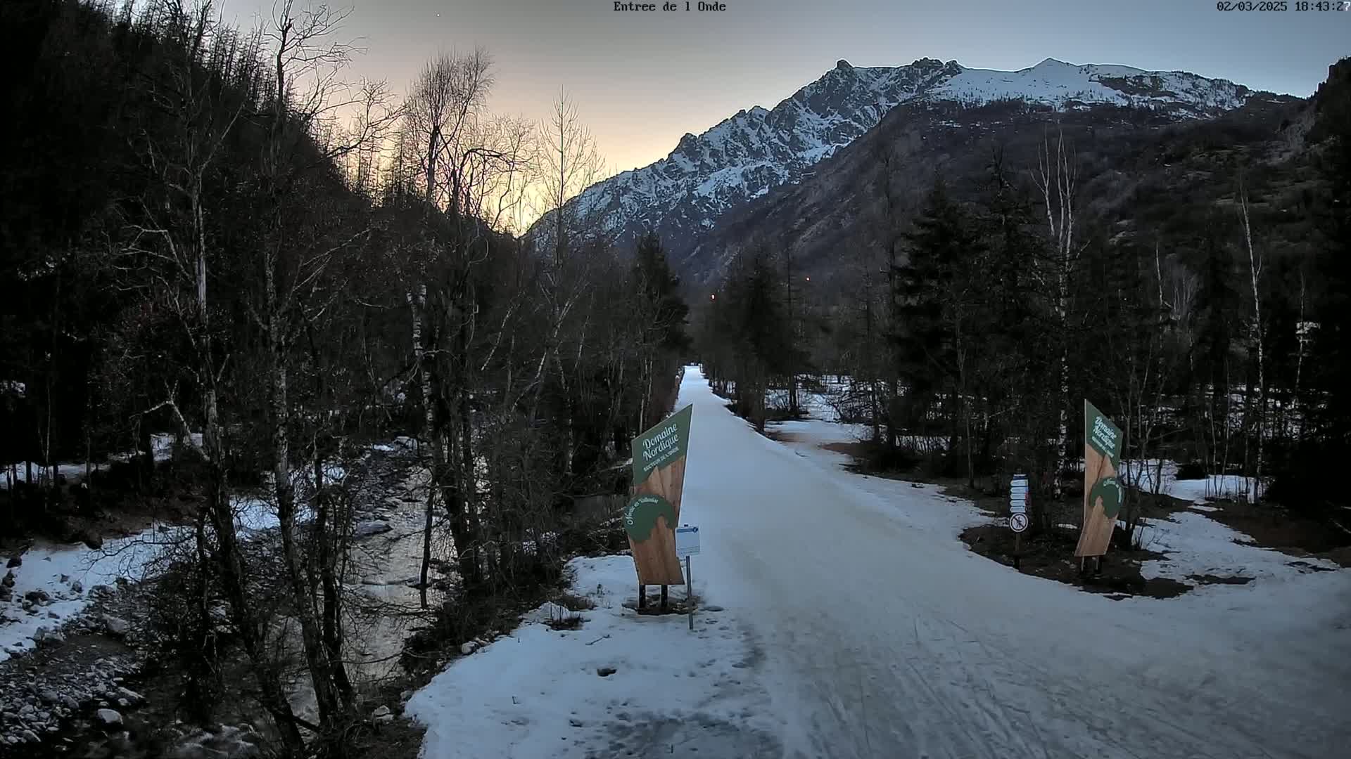 A snow-covered path winds through a valley at twilight, bordered by leafless trees and a snow-capped mountain range in the background.