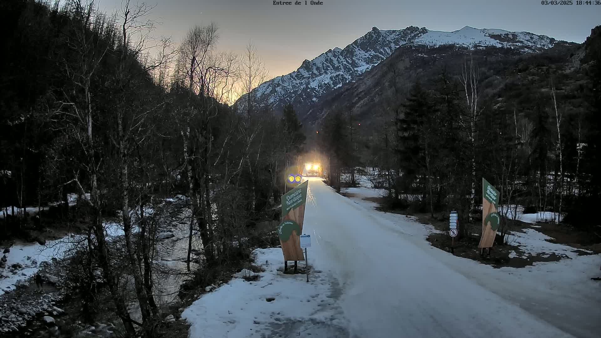 A snow-covered road winds through a valley at twilight, with a vehicle's headlights visible in the distance and snow-dusted trees lining the sides.