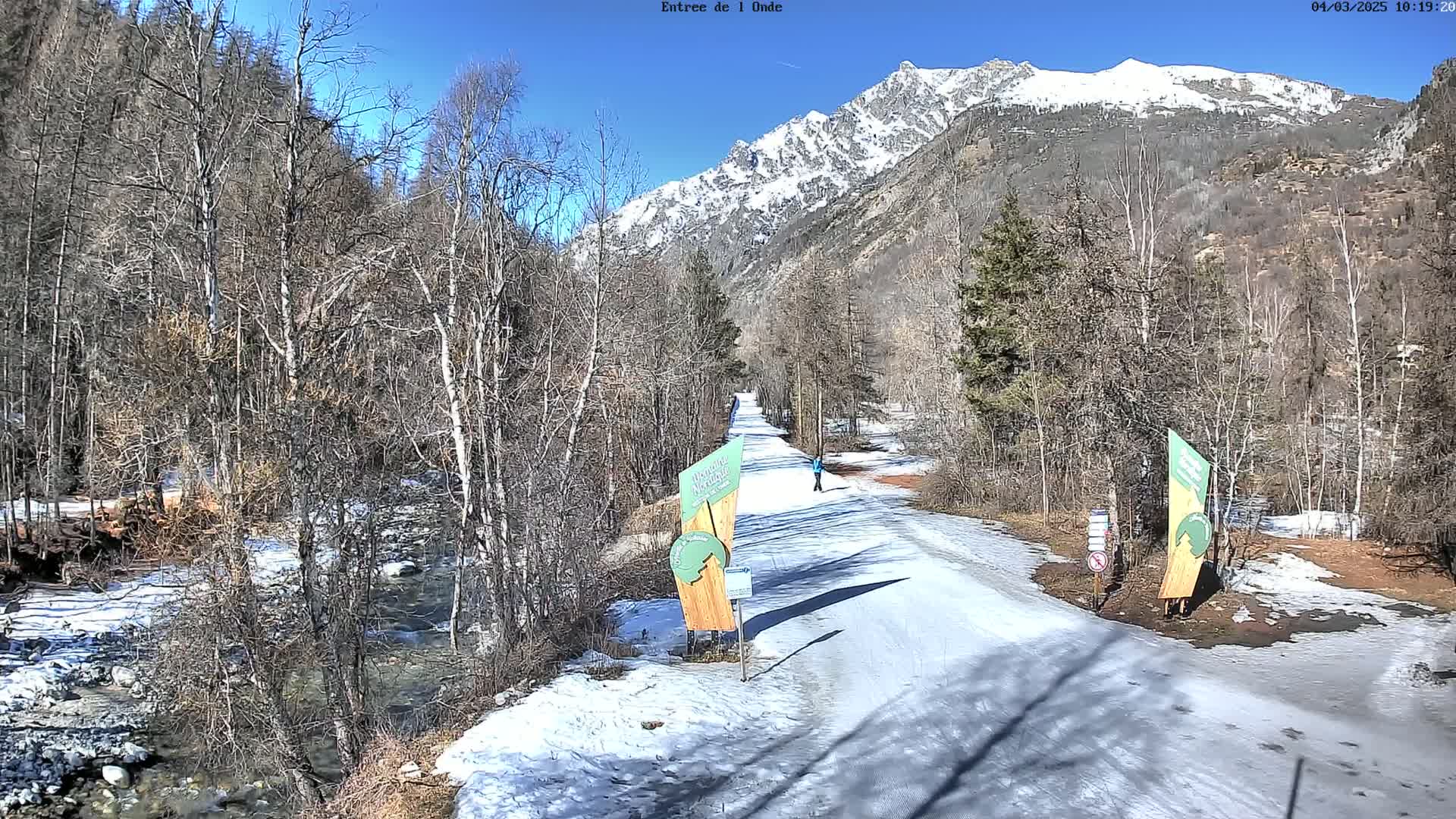 A snow-covered path winds through a valley with leafless trees and a snow-capped mountain in the background under a clear blue sky.