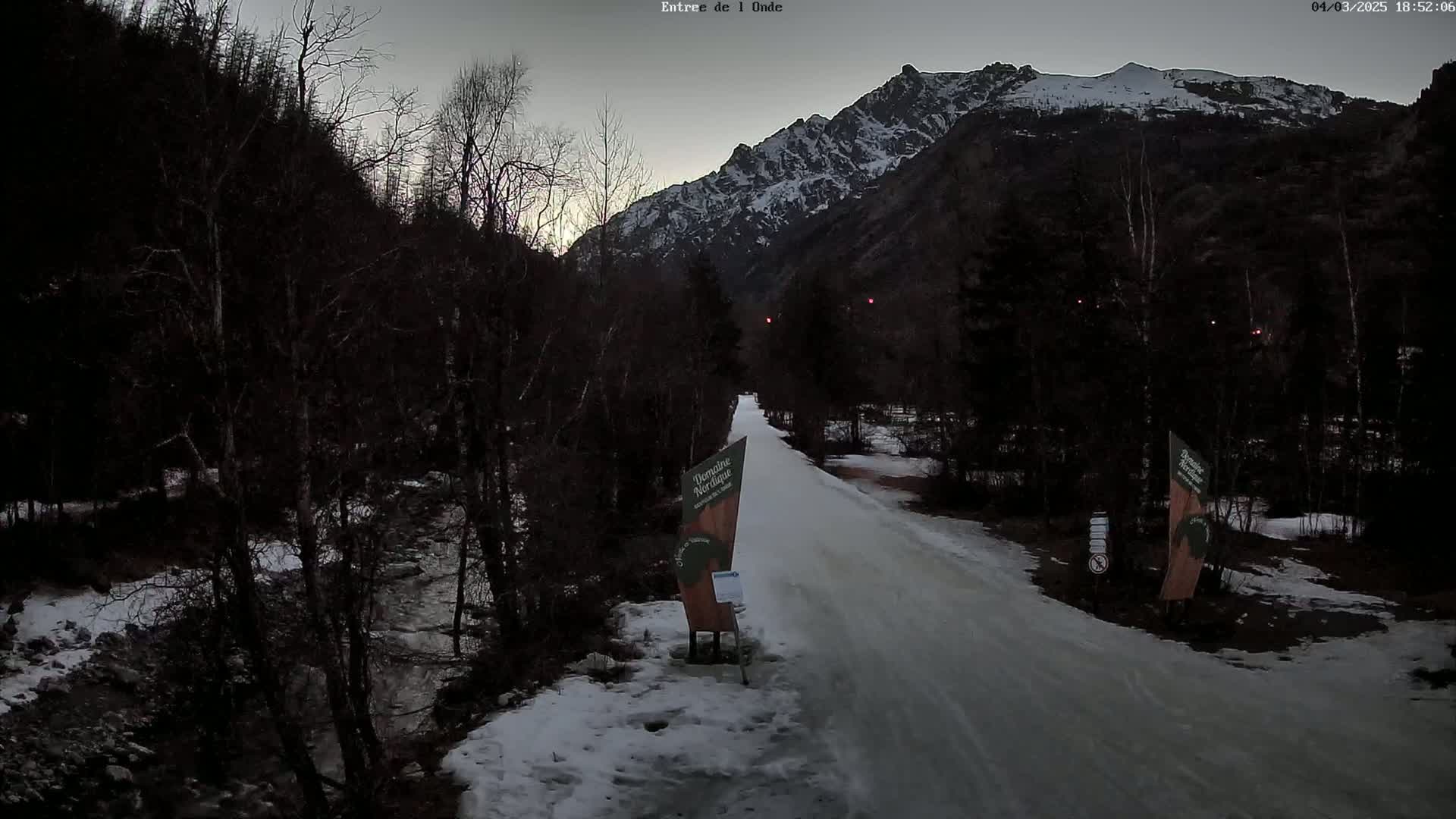A snow-covered road winds through a valley bordered by dark trees and snow-capped mountains under a dusky sky.
