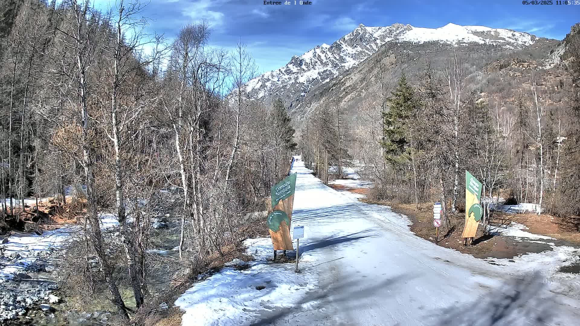 A snow-covered path winds through a valley of bare trees and a partly frozen stream, with a snow-capped mountain in the background under a sunny sky.