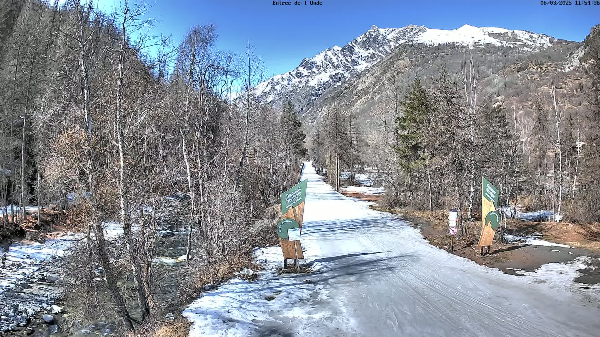 A snow-covered path winds through a valley of leafless trees towards a snow-capped mountain range under a clear blue sky.