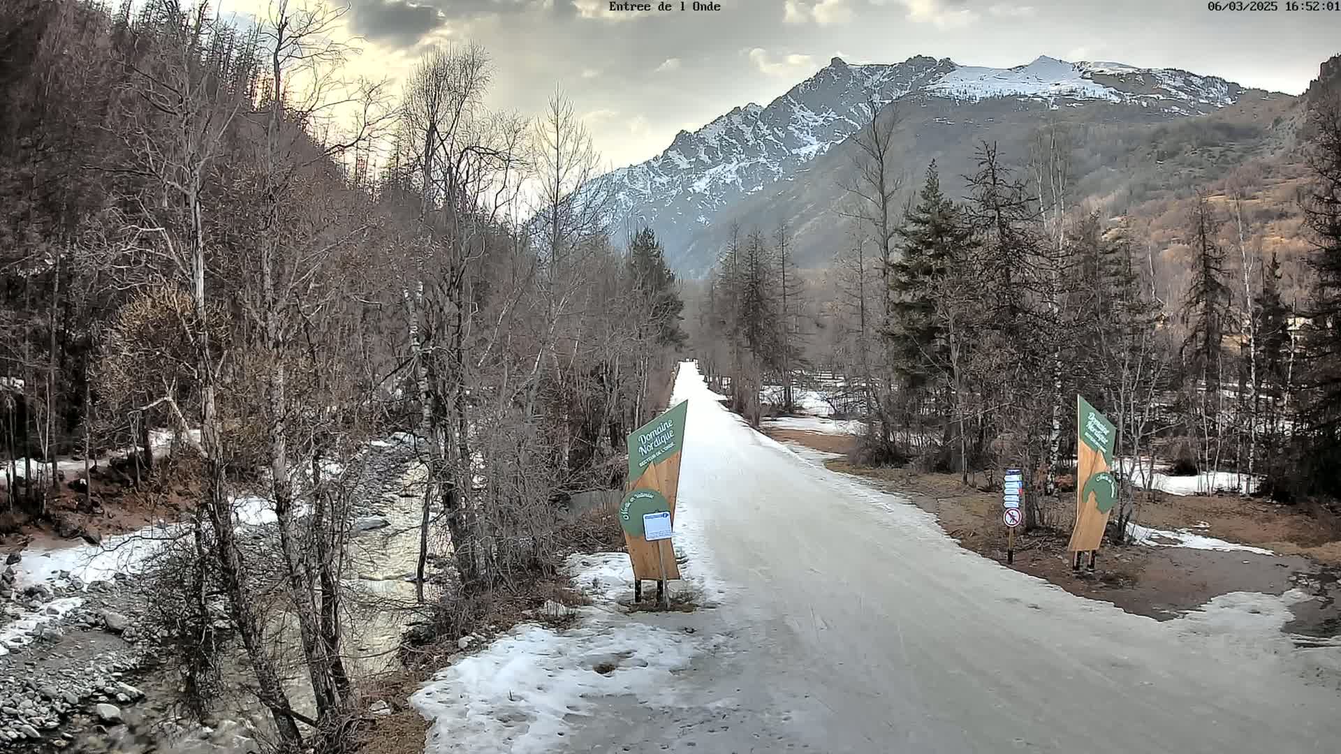 A snow-covered road leads toward a mountain range under a cloudy sky, with a stream running alongside it, and bare trees lining the path.