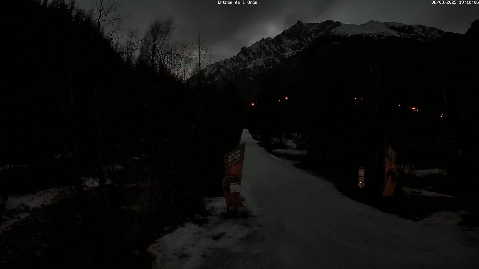 A snow-covered path winds through a dark, wooded area toward a snow-capped mountain under a dim, overcast sky.