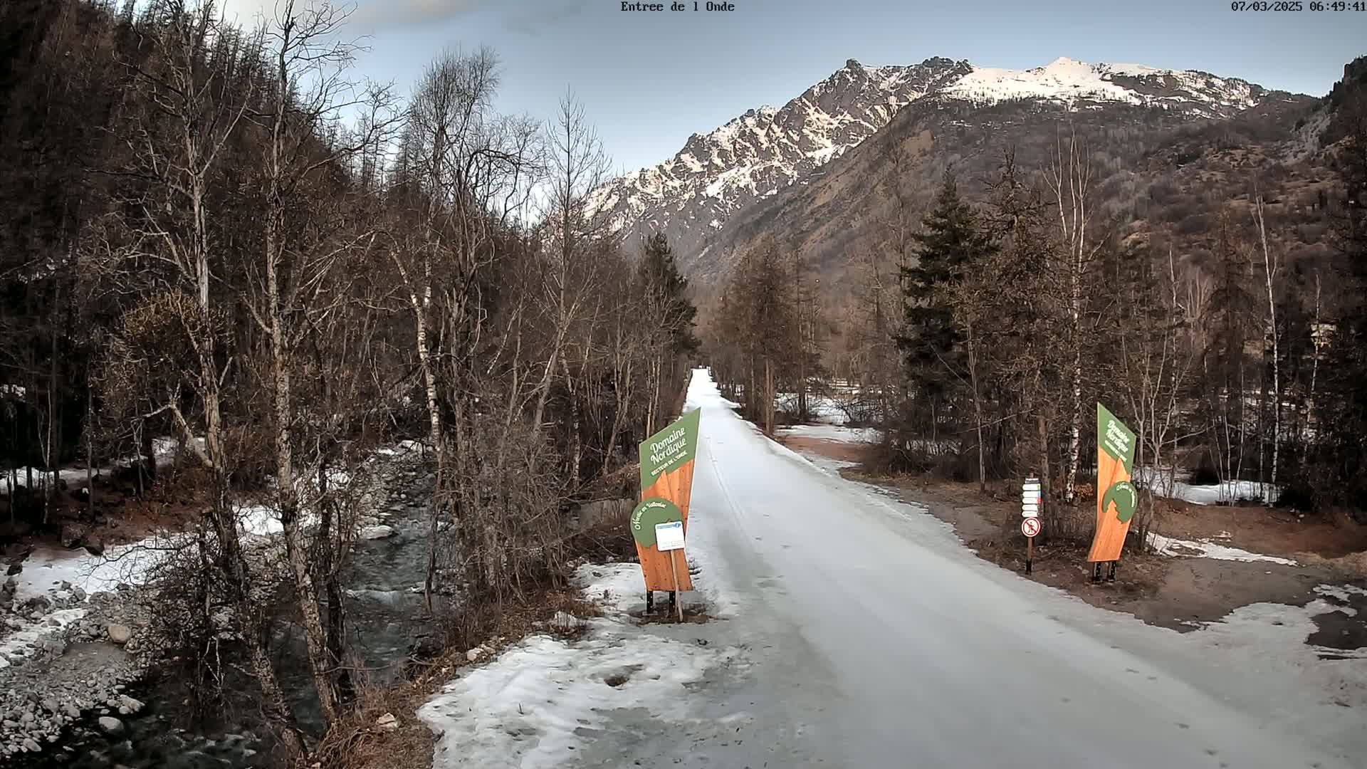 A snow-covered road winds through a valley of leafless trees toward a snow-capped mountain range under a mostly clear sky.