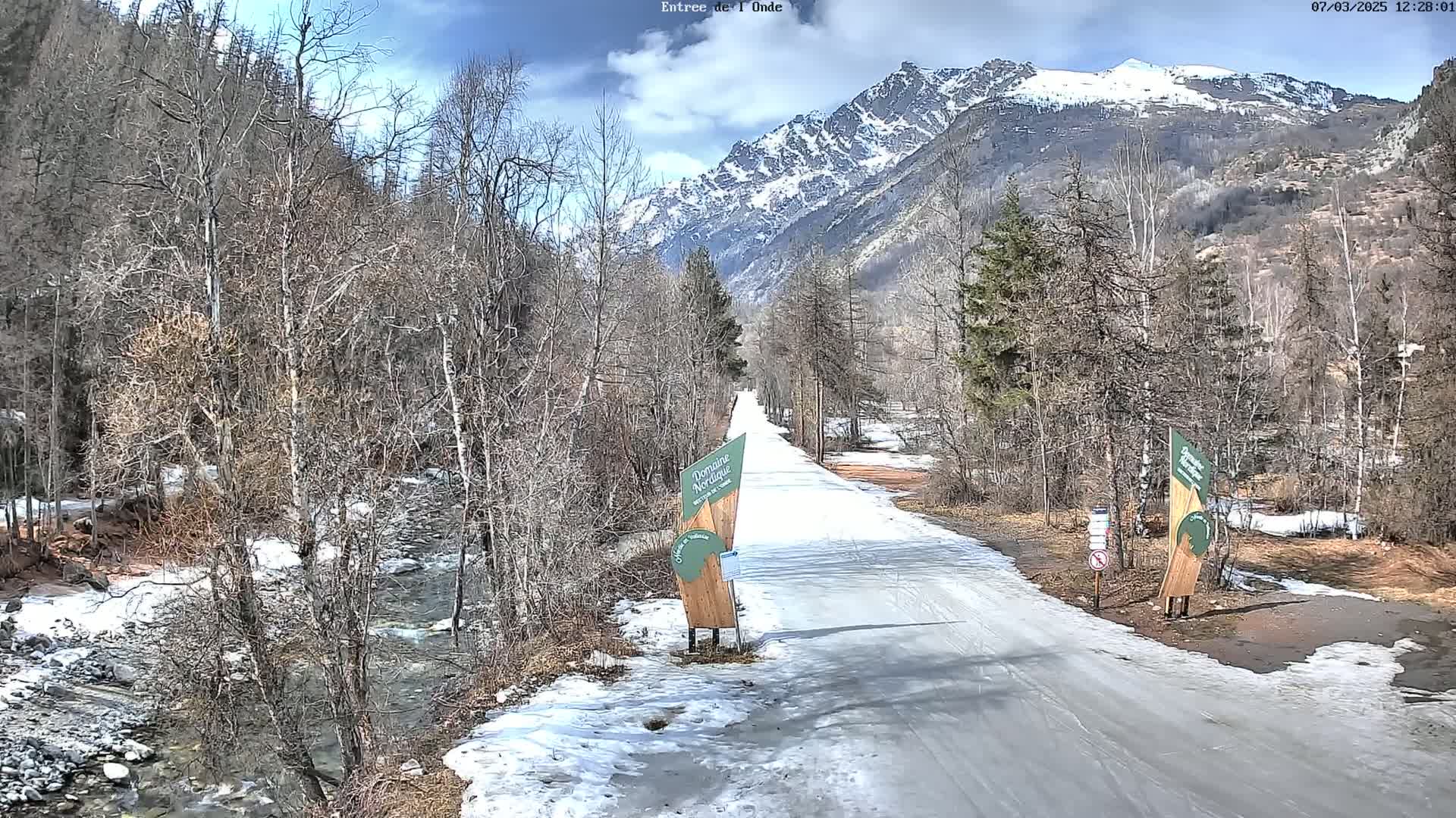 A snow-covered road winds through a valley of leafless trees towards a snow-capped mountain range under a partly cloudy sky.