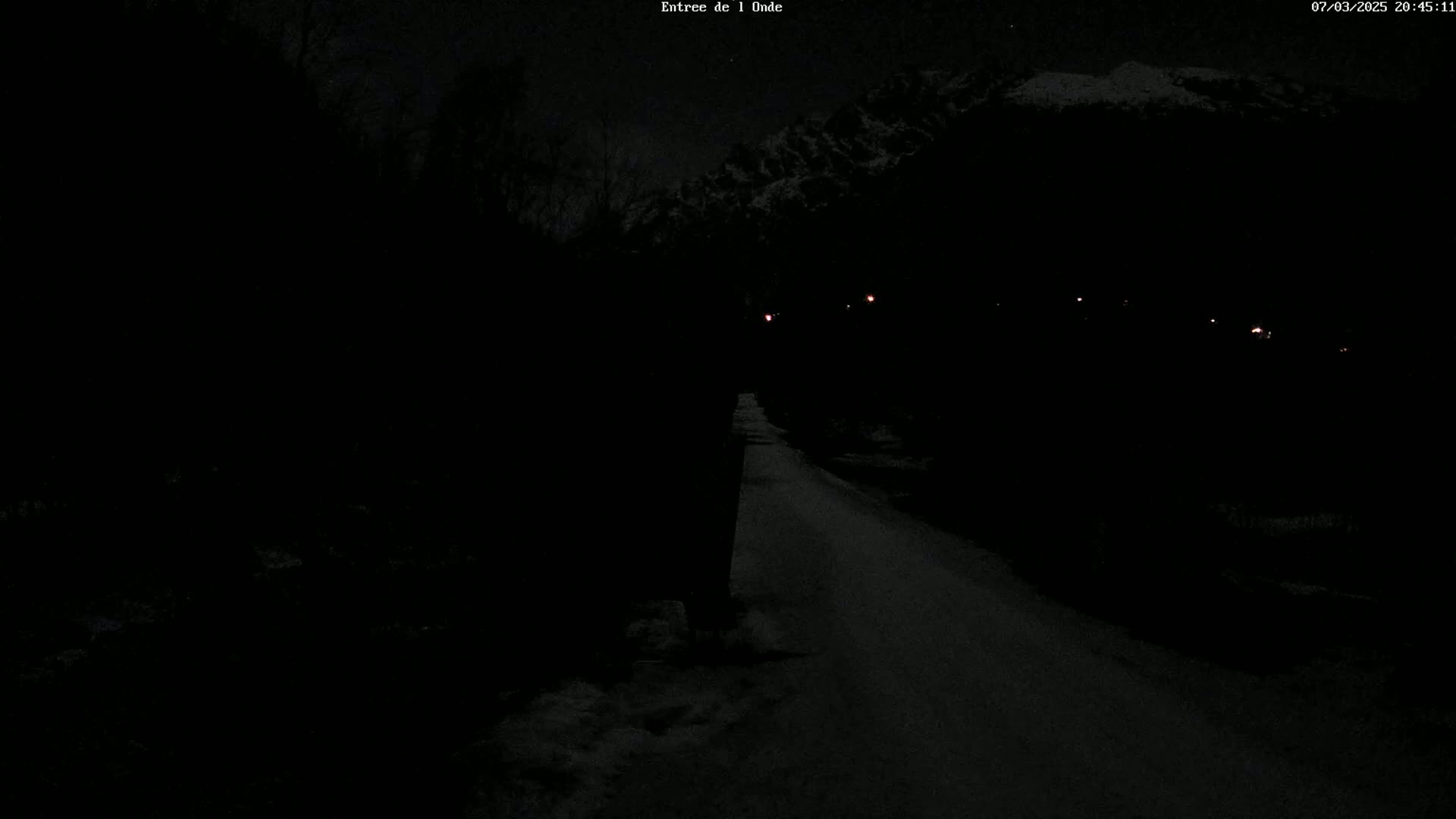 A dark, snow-covered road at night is illuminated by a few distant lights, with dark trees and snow-covered mountains in the background under a night sky.