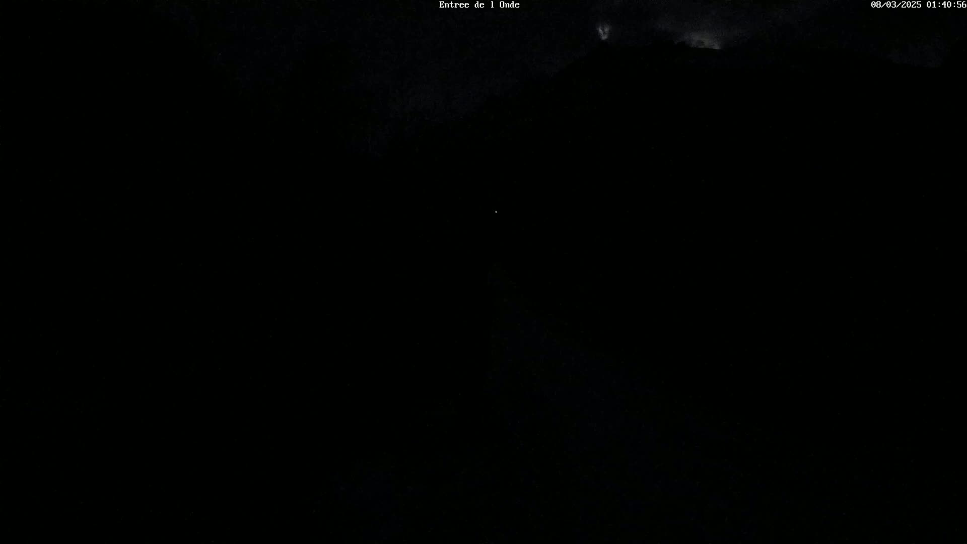 A dark, nighttime scene shows a dimly lit road leading towards a mountain range under a cloudy sky.