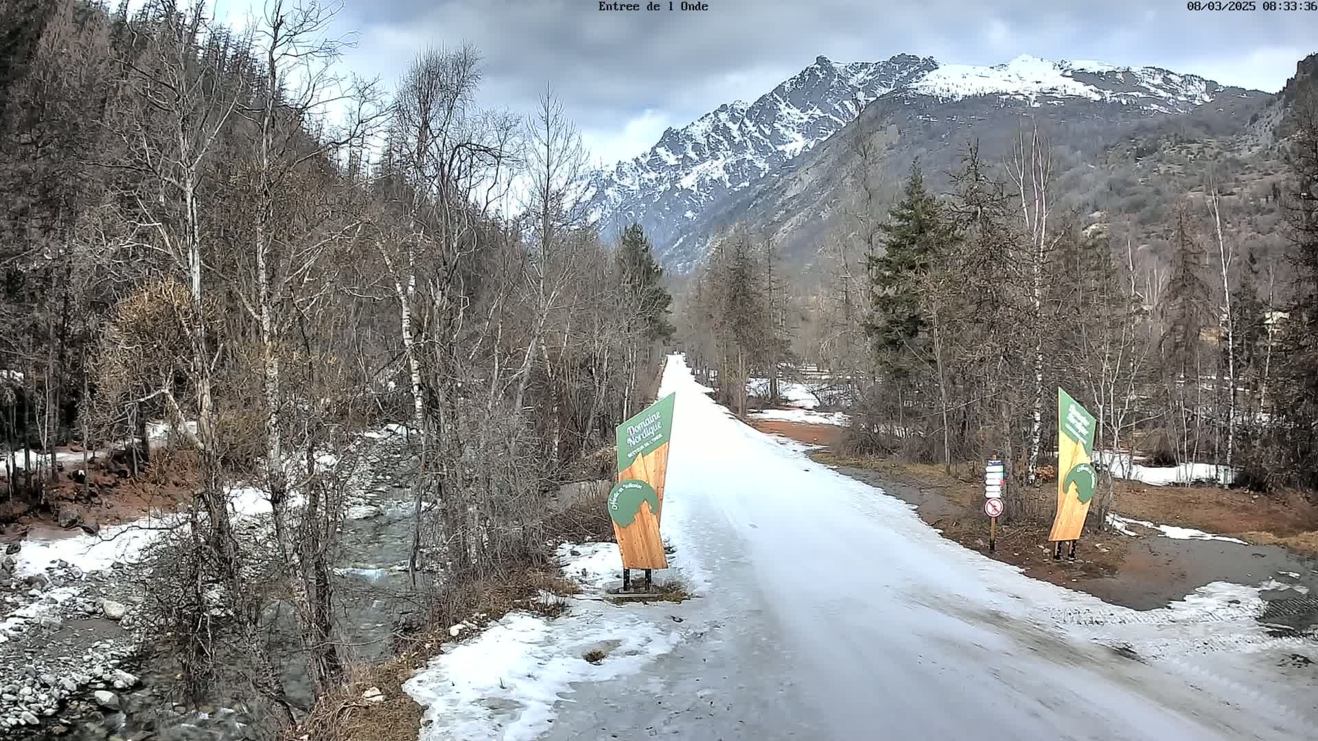 A snow-covered road winds through a valley of leafless trees and a partially snow-capped mountain range under an overcast sky, with a small stream visible alongside the road.