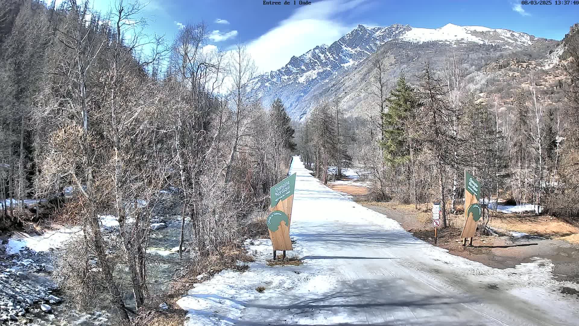 A snow-dusted road winds through a valley of leafless trees and a fast-flowing stream toward a snow-capped mountain range under a partly sunny sky.