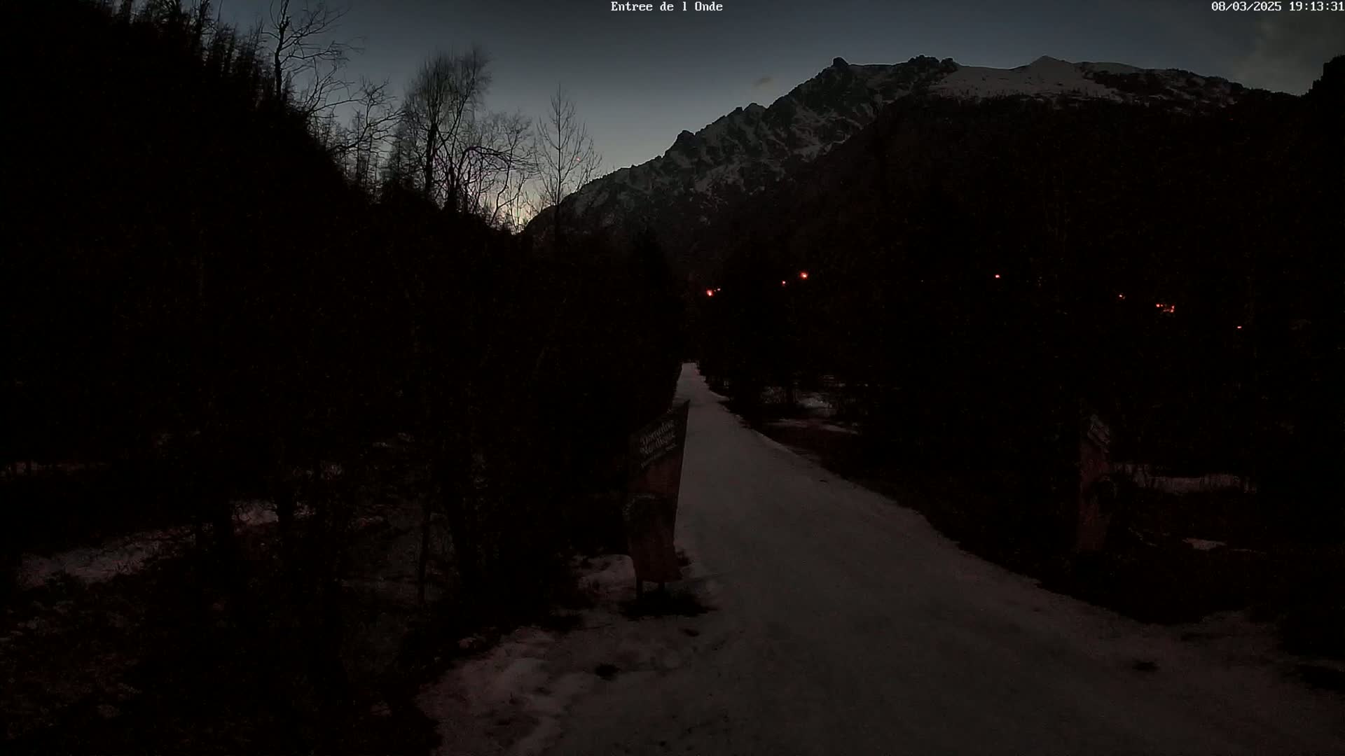 A snow-covered path winds through a dark, wooded area toward a snow-capped mountain under a twilight sky.