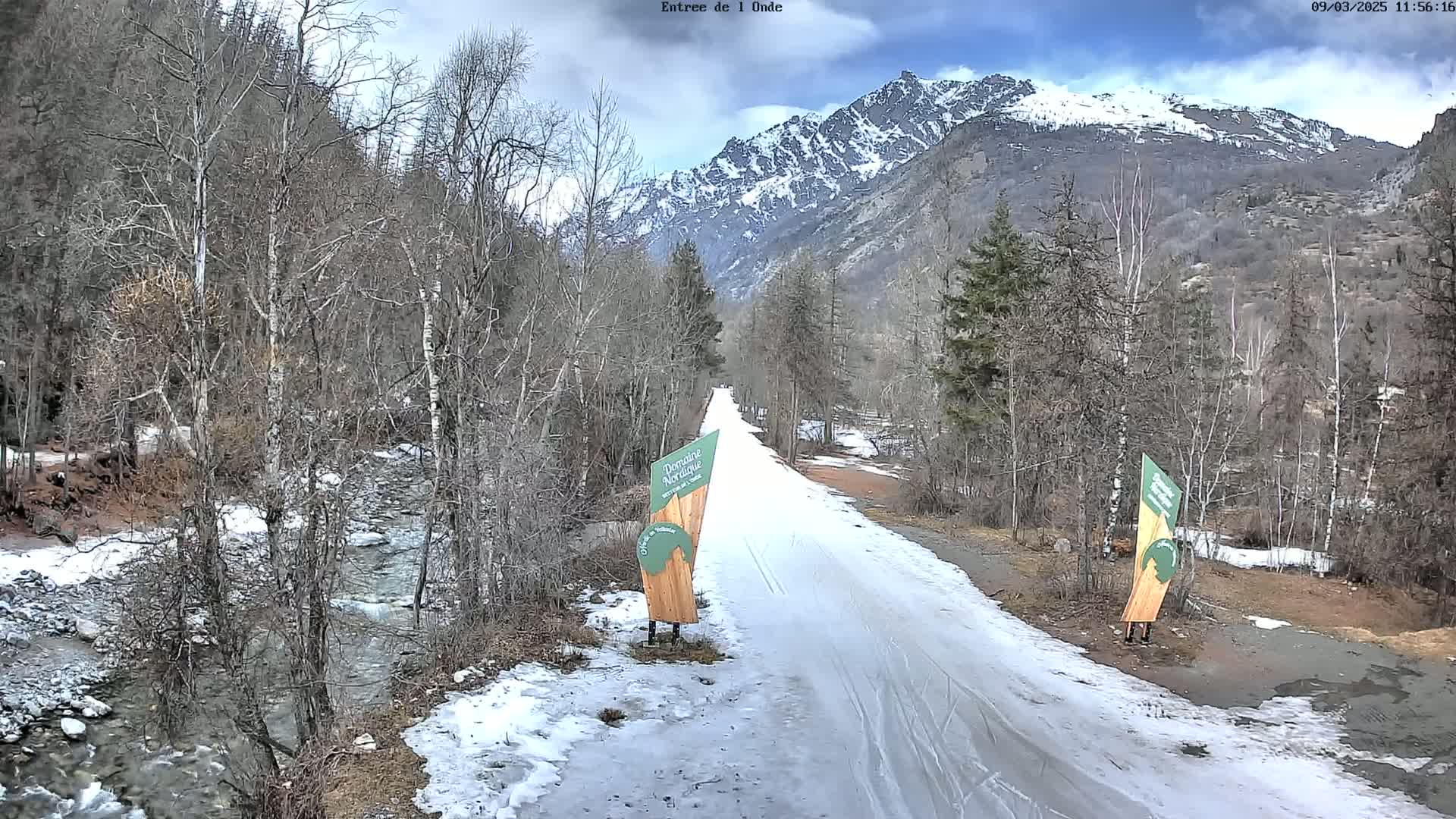 A snow-covered path winds through a valley of leafless trees alongside a partially frozen stream, with snow-capped mountains in the background under a partly cloudy sky.