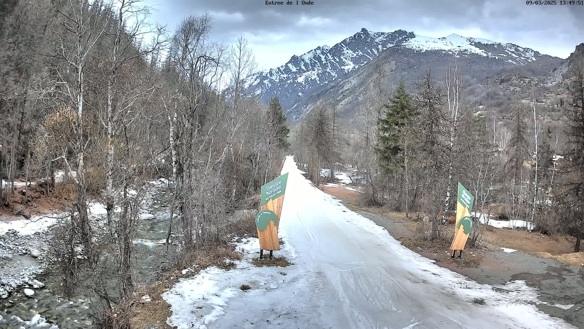 A snow-covered road winds through a valley, flanked by leafless trees and a partially snow-capped mountain range under a cloudy sky, with a small stream visible to the left.