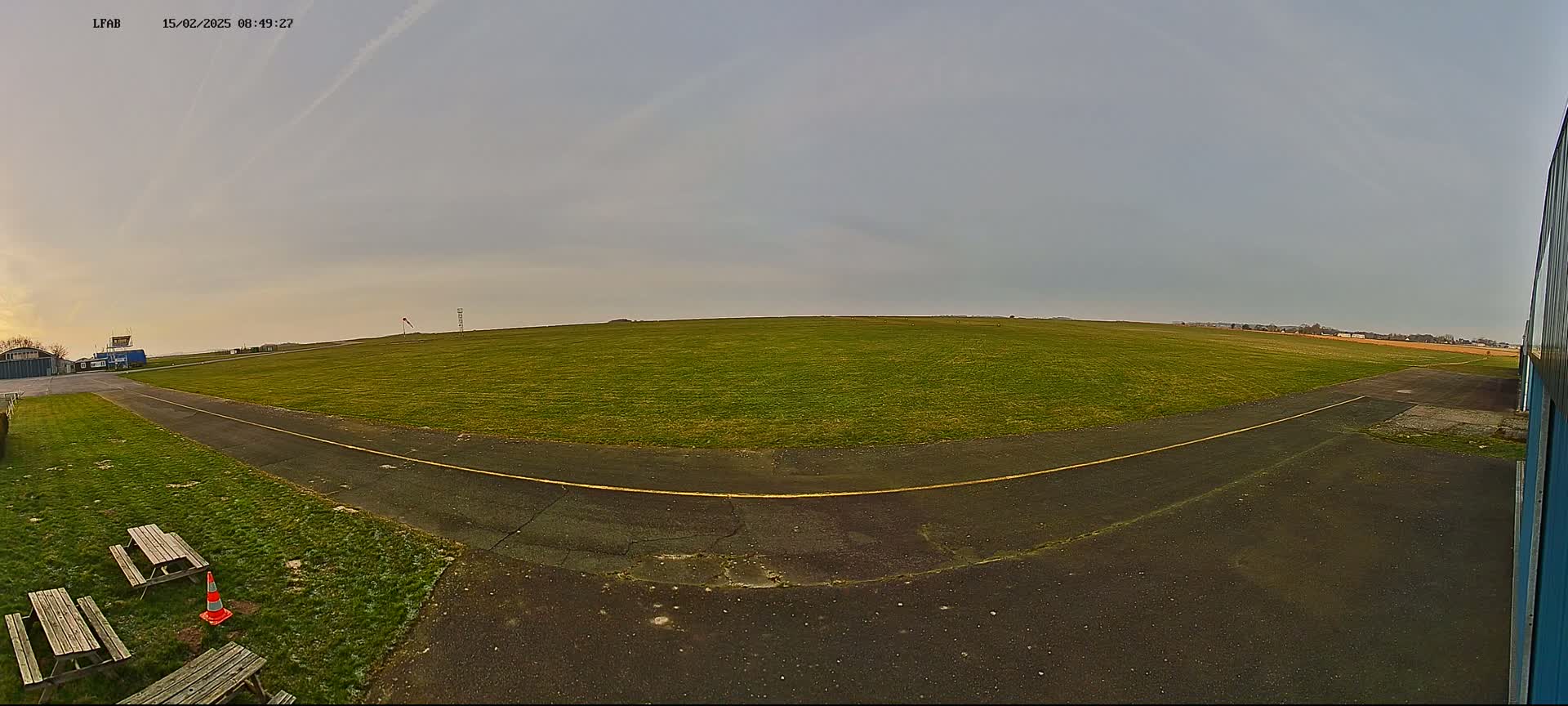 A grassy airfield with a paved taxiway and picnic tables is visible under an overcast sky.