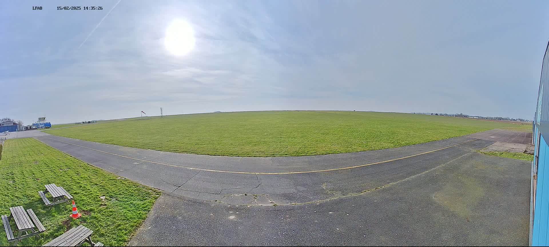 A sunny day at a grassy airfield shows a paved taxiway curving away from the viewer toward a distant hangar, with picnic tables and a traffic cone visible in the foreground.